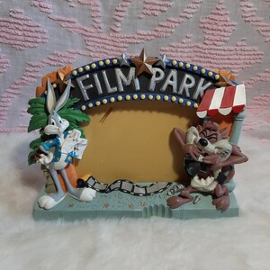 May include: A decorative photo frame featuring Bugs Bunny and the Tasmanian Devil. The frame is shaped like a movie set with the words "FILM PARK" across the top. The frame has a light blue base and a yellow center.
