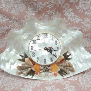 May include: A decorative clock with a white, shell-shaped frame. The clock face is white with black numerals and hands. The base is adorned with seashells and green accents, creating a coastal theme.