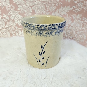 May include: A beige stoneware crock with a blue floral design around the rim and a stylized blue plant motif on the side. The crock has a cylindrical shape and is set against a soft, white background.