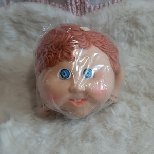 Puede incluir: Una cabeza de muñeca de plástico con pelo de lana rojo, ojos azules y una boca sonriente. La cabeza de la muñeca está envuelta en plástico.