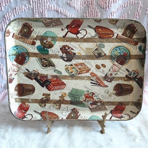 May include: A rectangular metal tray featuring a vintage-style collage of various items including bottles of Steinhäger, cigars, pipes, playing cards, dice, and chess pieces set against a background of printed newspaper text and horizontal stripes.