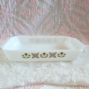 May include: A rectangular white milk glass baking dish with integrated side handles. The exterior features a repeating pattern of stylised green and yellow flowers and geometric leaf motifs arranged in a horizontal row along the centre.