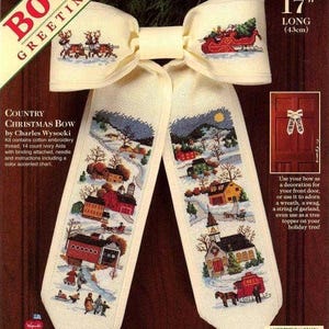 May include: A 43cm long ivory fabric front door bow featuring a counted cross stitch design by Charles Wysocki. The bow displays a winter village scene with houses, a church, a horse-drawn sleigh, and reindeer. The kit includes embroidery thread.