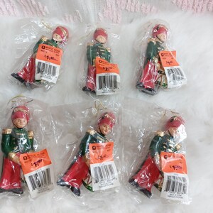May include: Six Christmas tree ornaments of toy soldiers in green jackets, red trousers, and red hats. The ornaments are packaged in clear plastic with gold string hangers. Each has a price tag.