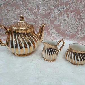 May include: A gold-coloured tea set with white vertical stripes. The set includes a teapot with a lid, a creamer, and a sugar bowl. The set is displayed on a white, fluffy surface.
