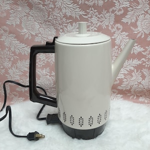 May include: A vintage cream-coloured electric coffee pot with a black handle, base, and power cord. The pot has a decorative black floral pattern around the base. The lid has a small, white knob. The coffee pot is plugged into an electrical outlet.