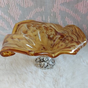May include: A brown and amber glass bowl with a swirl design and a silver metal base. The bowl is on a white fluffy surface.