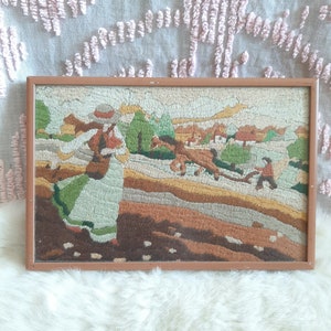 May include: A framed needlepoint picture of a woman in a green and white dress walking in a rural landscape. The scene includes a horse-drawn cart and other figures in the distance.