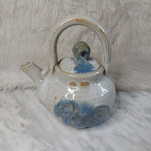 Peut inclure: Une petite théière en céramique blanche avec un glaçage bleu et un couvercle en forme de spirale. La théière a une poignée et un bec.