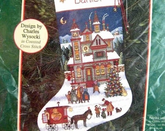 May include: A cross stitch stocking kit featuring a winter scene with a house, a Christmas tree, and a horse-drawn carriage. The stocking is white with a blue background and the name "Daniel" is embroidered at the top. The kit includes charms, beads, and instructions for counted cross stitch.