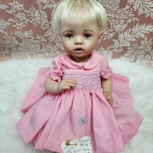 Op de afbeelding: Een porseleinen pop met blond haar en blauwe ogen, gekleed in een roze jurk met een blauw geborduurd lijfje en een vlinder. De pop heeft een label met de woorden "Sweet Thing" en andere handgeschreven details.