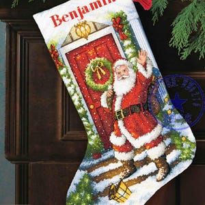 May include: A personalized Christmas stocking with a red door, a wreath, and Santa Claus holding a bag of gifts. The stocking is white with the name "Benjamin" embroidered in red.
