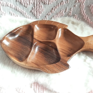 May include: A wooden serving tray shaped like a fish with four separate compartments. The tray is made of dark brown wood and has a natural, rustic finish.
