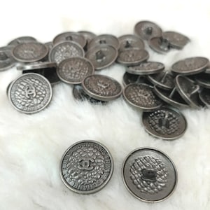May include: A collection of round, dark metallic buttons featuring a textured surface with a central interlocking CC logo. The outer rim of the buttons is inscribed with the text 31 RUE CAMBON PARIS FRANCE CHANEL.
