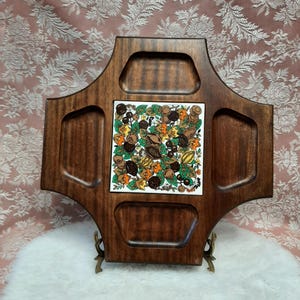 May include: A vintage wooden serving tray with four compartments and a central square tile insert. The tile features a colourful botanical design with nuts and leaves. The tray is supported by decorative metal feet.
