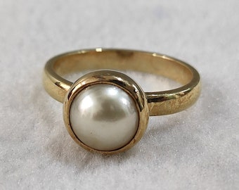 Könnte beinhalten: Ein goldener Ring mit einem weißen Perlmutt-Stein in einer Zargenfassung.