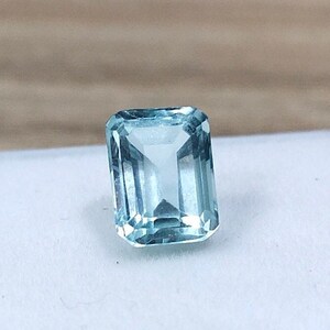 Puede incluir: Una piedra preciosa de aguamarina azul claro con talla esmeralda, expuesta en una caja blanca.