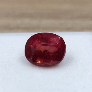 Könnte beinhalten: Ein ovaler Edelstein in tiefroter Farbe in einer weißen Schmuckschatulle.