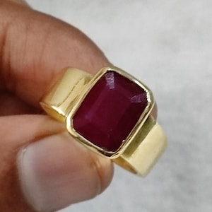 Könnte beinhalten: Ein goldfarbener Ring mit einem großen, rechteckigen, tiefroten Edelstein. Der Ring hat ein schlichtes, modernes Design mit einem dicken Band und einer Lünettenfassung, die den Stein sicher hält. Die Facetten des Edelsteins fangen das Licht ein.