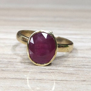 Könnte beinhalten: Ein goldener Ring mit einem großen, ovalen Rubin-Edelstein. Der Rubin ist tiefrot und hat einen facettierten Schliff.