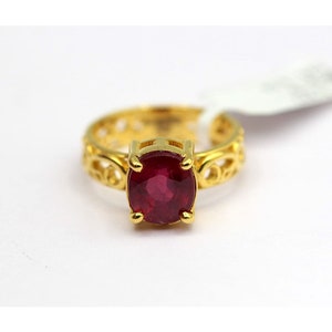May include: A gold ring featuring a deep red, oval gemstone. The ring's band has an intricate, filigree pattern. The gemstone is secured by four prongs. The ring is displayed against a white backdrop.