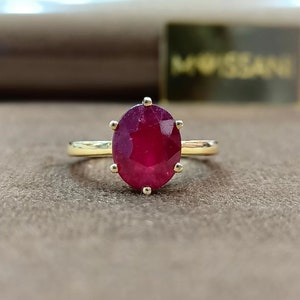 Puede incluir: Un anillo de oro con una gran gema de rubí de corte ovalado. El rubí es de color rojo intenso y está sujeto por seis garras. La banda del anillo es de oro pulido. El fondo es de color marrón neutro.
