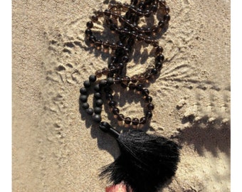 May include: A brown gemstone mala necklace with a black tassel. The necklace is made of round beads and is lying on a sandy surface.