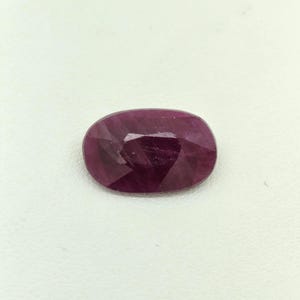 Peut inclure: Une pierre précieuse rubis de forme ovale avec une teinte rouge magenta profond et une surface à facettes. La pierre présente des inclusions internes et une finition polie, reflétant la lumière sur ses multiples facettes taillées.