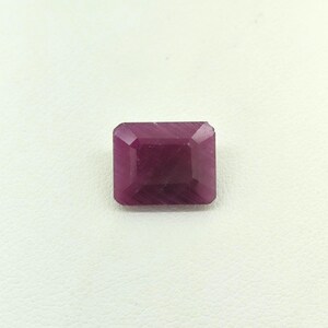 May include: A rectangular, faceted gemstone with a deep magenta-red colour. The stone features a step-cut design with clear, geometric edges and a polished surface that reflects light across its flat top and angled side facets.