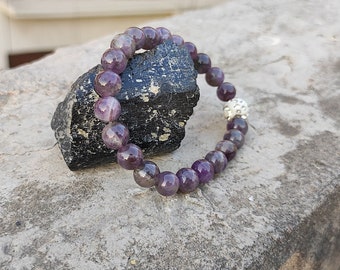 May include: A purple amethyst gemstone bracelet with a silver bead charm. The bracelet is resting on a black stone.