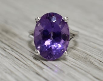 May include: A silver ring with a large, oval-shaped amethyst gemstone. The gemstone is a deep purple color and has a faceted cut.