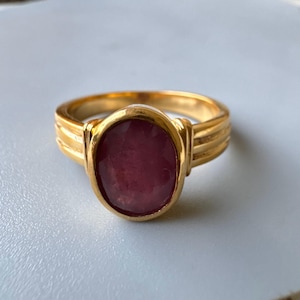 Könnte beinhalten: Ein goldener Ring mit einem tief roten ovalen Edelstein, der in einer Zargenfassung sitzt. Der Ringarm hat ein strukturiertes Design.