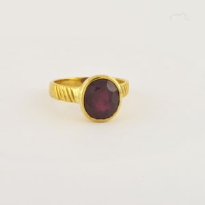 May include: A gold ring with a deep red oval gemstone set in a bezel setting. The band of the ring has a twisted design.