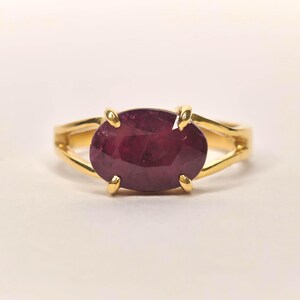 Op de afbeelding: Een gouden ring met een grote, ovaalvormige, diepmagenta edelsteen in een zetting met vier pootjes. De ringband is gespleten in twee dunne, gepolijste gouden segmenten die aan de zijkanten samenkomen, wat zorgt voor een strak en open ontwerp.