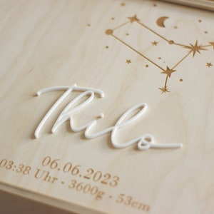 Könnte beinhalten: Eine Holzkiste mit einer weißen Inschrift des Namens "Thilo" und dem Datum "06.06.2023" sowie der Uhrzeit "03:38 Uhr" und den Maßen "3600g - 53cm". Die Kiste ist mit einem Sternbild-Design verziert.