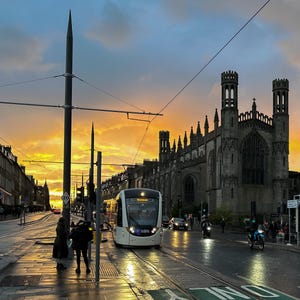 Puede incluir: Un tranvía blanco con la palabra "Trans" en la parte delantera viaja por una calle mojada frente a un gran edificio de piedra. El cielo es una mezcla de naranja y azul. La gente está en la acera. Se ven farolas y cables aéreos.