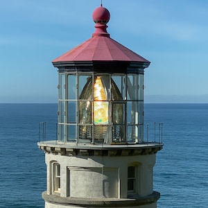 Puede incluir: Un faro blanco con un techo rojo y una lente de vidrio en la parte superior. El faro está en un acantilado con vistas al océano.