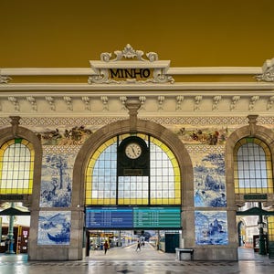 Puede incluir: Vista interior de una estación de tren con detalles arquitectónicos ornamentados. El techo es amarillo y las paredes presentan murales de azulejos azules y blancos. Un gran reloj está centrado sobre la entrada, con la palabra "MINHO" encima.