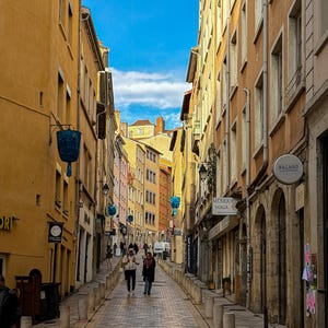 May include: A cobbled street in a European town, flanked by buildings in shades of yellow, orange, and pink. The street leads towards the horizon under a blue sky. People are walking along the street. Street signs and blue lanterns hang from the buildings.