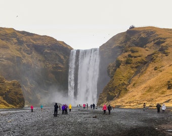 Könnte beinhalten: Ein großer Wasserfall stürzt von einer felsigen Klippe herab, mit einer Gruppe von Menschen, die am Fuße stehen. Der Wasserfall ist umgeben von grünen Hügeln und einem bewölkten Himmel.