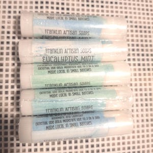 May include: A stack of five white lip balm tubes with labels. The labels read "Franklin Artisan Soaps" and list various scents, including "Eucalyptus Mint" and "Rosemary, and Lavender". The text "Made Local in Small Batches" is also visible.