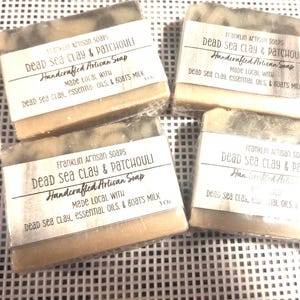 Könnte beinhalten: Vier handwerkliche Seifenstücke mit Etiketten mit der Aufschrift "Dead Sea Clay & Patchouli". Die Seife ist hellbraun mit einer dunkleren grauen Oberseite. Auf den Etiketten steht außerdem, dass die Seife handgefertigt ist mit Totes-Meer-Ton, ätherischen Ölen und Ziegenmilch.