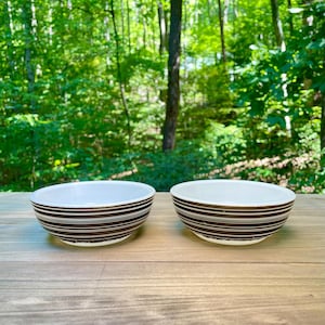May include: Two ceramic bowls with a white interior and a brown striped exterior. The bowls are round and sit on a wooden surface. The background is a blurred view of green trees.