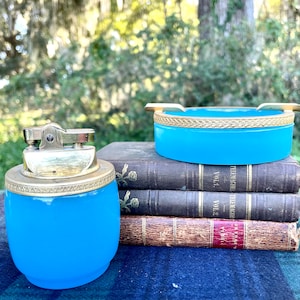 May include: A vintage blue glass ashtray and lighter set with gold trim. The set is sitting on top of a stack of old books.