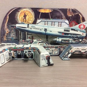 May include: A detailed papercraft model of a futuristic spaceship and control room. The spaceship is silver with red accents and a distinctive tail fin. The control room features intricate details and a figure in a circular window.