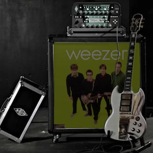 May include: A black guitar amplifier cabinet featuring the Weezer green album cover art on its front panel, topped with a black electronic head unit, and accompanied by a white electric guitar and a smaller black equipment case on the side.