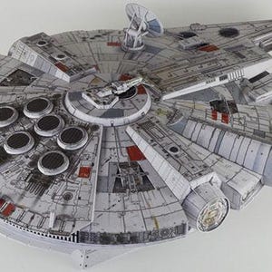 May include: A detailed model of the Millennium Falcon starship, featuring a gray and white color scheme with red accents. The model has a circular main body with multiple circular vents and detailed paneling, and two forward mandibles.