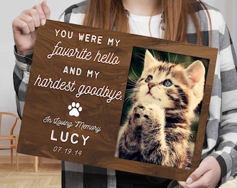 May include: A rectangular wooden memorial plaque featuring a photo of a tabby kitten. The text reads: YOU WERE MY favorite hello AND MY hardest goodbye. In Loving Memory LUCY 07.19.14, accompanied by a white paw print icon on the dark wood grain surface.