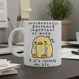 Op de afbeelding: Witte keramische mok met een humoristisch ontwerp. De mok heeft een huilende gele kuiken cartoon met de tekst "accidentally became important at work... & it's ruining my life." De mok staat op een donkerbruin bureau met een laptop en kantoorbenodigdheden.