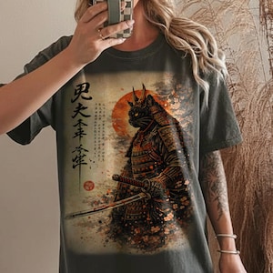 May include: Dark gray t-shirt featuring a detailed illustration of a black cat samurai with a sword, set against a rising sun and Japanese text. The design incorporates elements of traditional Japanese art.